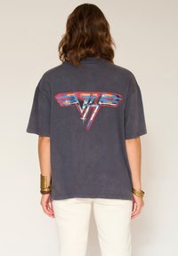 Personne avec des cheveux bruns ondulés de dos, portant un t-shirt noir oversized avec un graphique vintage coloré dans le dos et un pantalon blanc.