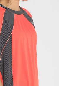 Krátký rukáv sportovní tričko v jasně korálové barvě s černými síťovinovými akcenty a švy. Má kulatý výstřih a volný střih.