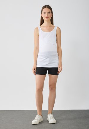 Weißes Tanktop mit rundem Ausschnitt und figurbetontem Schnitt, kombiniert mit schwarzen Shorts. Das Modell trägt weiße Sneakers. Glatter Stoff und lässiges Design.