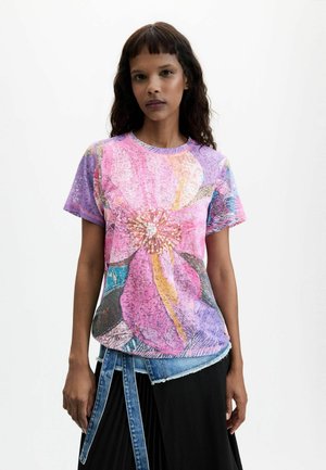 Kobieta o średniej długości ciemnych włosach nosi różowo-fioletowy t-shirt w kwiaty oraz czarną spódnicę z detalem z dżinsowym paskiem.