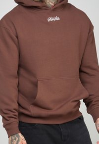 Felpa con cappuccio marrone realizzata in tessuto morbido, con tasca frontale, polsini a coste e un piccolo logo bianco sul petto.