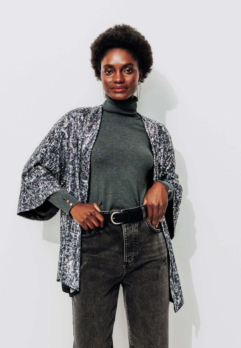 Un kimono à motifs noir et blanc sur un col roulé gris, associé à un jean en denim taille haute. Il présente des manches larges et une ceinture.