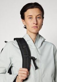 Helles graues Zip-Jackett mit Stehkragen. Verfügt über einen schwarzen, strukturierten Rucksacktrageriemen und verstellbaren Befestigungselementen. Glattes, elegantes Material.