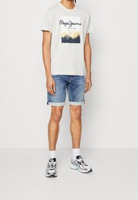 Miesmalli, joka pukeutuu vaaleanharmaaseen Pepe Jeans London -t-paidan, käärittyihin farkkushortseihin, valkoisiin sukkiin ja hopeisiin Asics-juoksukenkiin, seisoo yksinkertaisella taustalla.