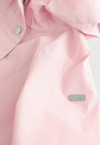 Giacca impermeabile rosa chiaro con texture liscia, chiusura a scatto e dettaglio logo argentato sul lato sinistro.
