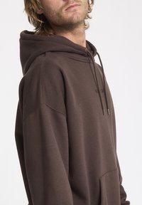 Sweat-shirt à capuche marron en tissu doux, doté d'une poche kangourou et d'une fermeture à cordon. Marquage subtil sur la poitrine.