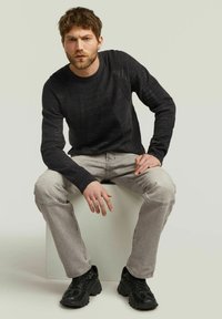 Pull en tricot noir à côtes avec un col ras du cou, associé à un pantalon gris clair, assis sur un bloc blanc, portant des baskets noires.