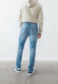 Jeans de denim azul claro con corte slim, bolsillos traseros tipo parche y costuras sutiles, combinados con una sudadera beige y zapatillas oscuras.