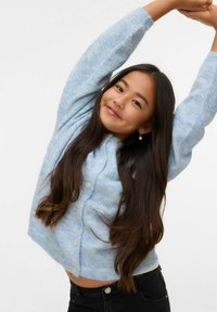 Šviesiai mėlynas trumpas megztinis su minkšta tekstūra, apvali apykakle, priekyje sagos ir ilgomis rankovėmis, derinamas su juodais džinsais.