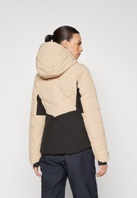 Padded Jacke mit einem Beigen und Schwarzen Farbblock-Design, Kapuze und einem Zickzackmuster auf der Rückseite, mit einer weichen, strukturierten Oberfläche.