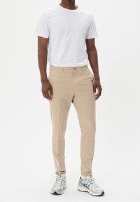 Pantalones beige de corte recto, con un cierre de un solo botón y bolsillos delanteros, combinados con una camiseta blanca básica y zapatillas deportivas.