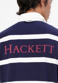 Polo bleu marine avec des rayures blanches, comportant un grand logo "HACKETT" en bordeaux à l'arrière. Matière en coton avec un design de col classique.