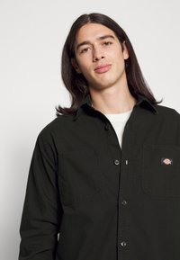 Camisa de botonadura en verde oscuro con dos bolsillos frontales, una textura suave y un pequeño parche con la marca en el bolsillo izquierdo.
