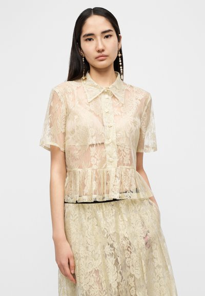 Δαντελένιο μπλουζάκι σε απαλό κίτρινο χρώμα με γιακά, κοντά μανίκια και κούμπωμα με κουμπιά. Σχεδιασμός πεπλούμ και διαφανές λουλουδάτο μοτίβο.