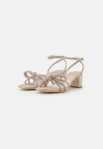 Sandales dorées à talons featuring un design noué avec des sangles ornées d'accents métalliques. Semelle texturée et talon épais pour un meilleur soutien.