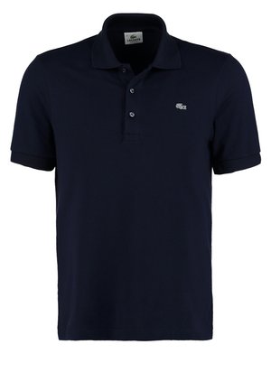 Baarddragende man die loopt, gekleed in een marineblauw poloshirt, blauwe broek en witte sneakers tegen een eenvoudige witte achtergrond.