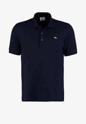 Baarddragende man die loopt, gekleed in een marineblauw poloshirt, blauwe broek en witte sneakers tegen een eenvoudige witte achtergrond.