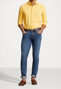 Hombre que lleva una camisa amarilla de manga larga y botones, jeans azules con los puños doblados y cinturón marrón, mocasines de piel marrón, y reloj de pulsera, con las manos en los bolsillos.