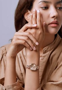 Reloj de pulsera de oro rosado con esfera cuadrada, correa de malla metálica y un dial de color claro que presenta marcadores minimalistas y una textura sutil.