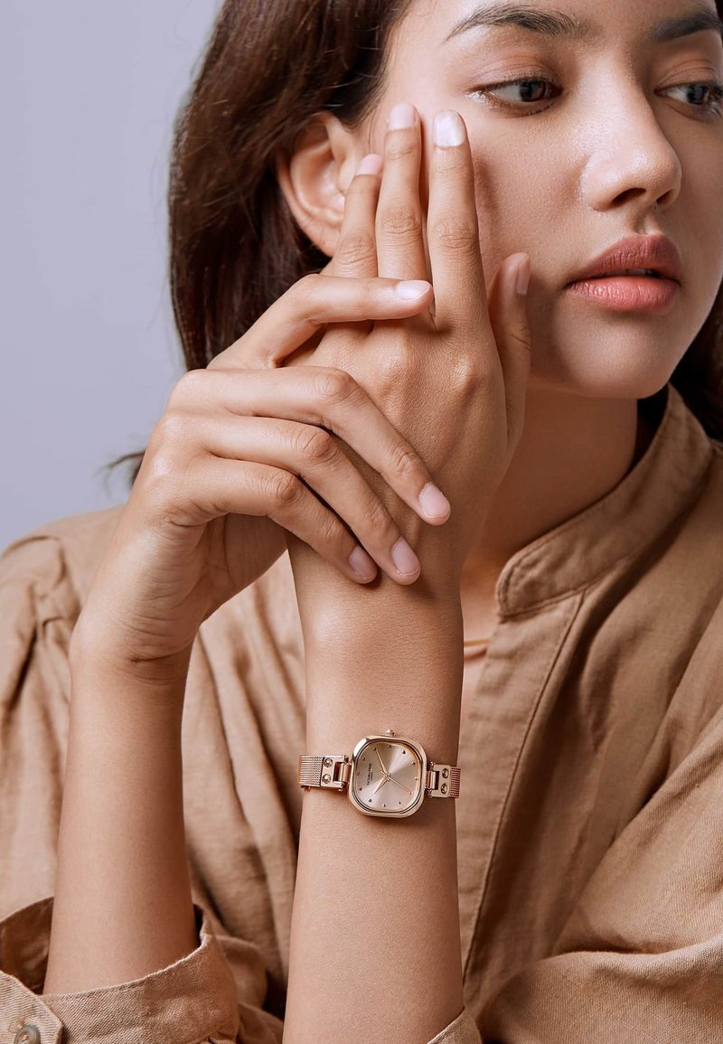Vrouw in beige overhemd met een gouden horloge met vierkante wijzerplaat, houdt de ene hand met de andere dicht bij haar gezicht en kijkt opzij.
