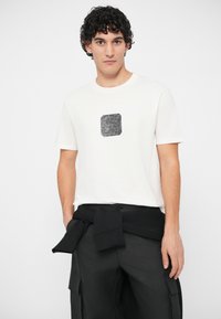 Homem com cabelo encaracolado a usar uma t-shirt branca com um gráfico quadrado preto e calças cargo pretas, sweatshirt preta atada à cintura, de pé.