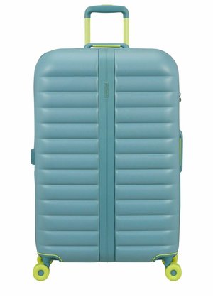 Grande valise rigide bleue avec des rainures horizontales, poignée jaune-vert et quatre roues pivotantes jaune-vert.