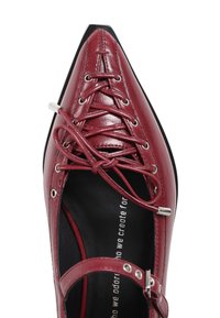 Zapato de cuero burdeos con punta afilada y diseño de cordones. Presenta ojales plateados y un forro interior negro con texto. Suela plana.