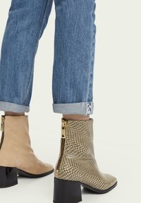 Botines: uno en gamuza beige, otro con un estampado de serpiente en beige y negro, ambos con cremallera dorada y tacones de bloque, combinados con denim enrollado.