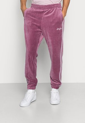 Mauve welurowe spodnie dresowe z białymi paskami po bokach, elastycznym paskiem w talii i ściągaczami. Połączone z białymi sneakersami dla kontrastu.
