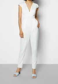 Mono blanco con escote en V profundo, mangas cortas y piernas ajustadas. Combina con mules abiertos en punta de color azul claro. Tela suave, diseño minimalista.
