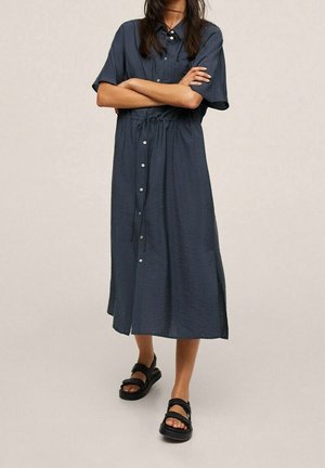 Kobieta stojąca z założonymi skrzyżowanymi ramionami, ubrana w midi sukienkę zapinaną na guziki w ciemnoniebieskim kolorze oraz czarne sandały z odkrytymi palcami na jasnym tle.