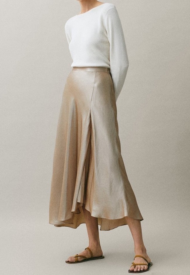 Vrouw in een witte langemouwen top, een beige satijnen midi-jurk met een open voorkant en tan platte sandalen, staande tegen een effen achtergrond.