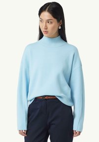 Hellblauer Rollkragenpullover aus weichem Material, mit langen Ärmeln und lockerer Passform. Getragen mit dunkelblauen Hosen und einem braunen Gürtel.
