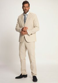 Beige Leinenanzug mit taillierter Passform, ein-knopf Blazer, gerade geschnittene Hose und einer gemusterten Krawatte. Akzentuiert mit schwarzen Slippern.