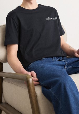 Osoba ubrana w czarny t-shirt z logo Gant 1949 i niebieskie dżinsy, siedząca na beżowym fotelu z drewnianymi podłokietnikami.