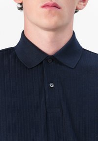Polo shirt azul marinho com textura canelada e colarinho abotoado, apresentando um pequeno bolso e um único botão de fecho. Textura suave e design clássico.