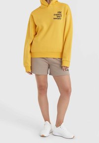 Żółta bluza z kapturem z logo z przodu, w zestawie z beżowymi szortami. Model ma na sobie białe tenisówki. Gładka faktura, luźny krój, casualowy design.