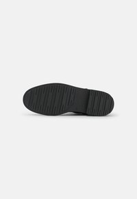 Suela de zapato de goma negra con ranuras para agarre, que presenta un diseño plano y detalles de marca sutilmente grabados cerca del área del talón.