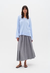 Helles blaues langärmliges Oberteil mit V-Ausschnitt, kombiniert mit einem grauen, gefalteten Maxirock. Das Outfit wird durch schwarze Flip-Flops ergänzt.