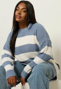 Niebiesko-biała sweter w paski o teksturowanym dzianinie, z okrągłym dekoltem, opadającymi ramionami i długimi rękawami. Połączony z dżinsowymi spodniami.