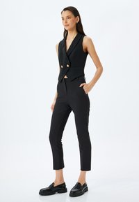 Zwarte op maat gemaakte vest met dubbele gouden knopen, gecombineerd met slim-fit zwarte broek en zwarte loafers. Gladde stof, slanke silhouette.