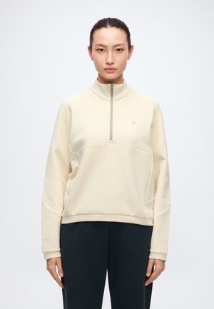 Femme avec les cheveux attachés, portant un pull beige clair à fermeture éclair quart et un pantalon noir, debout devant un fond blanc uni.