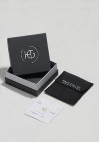 Juoda papuošalų dėžutė su pilku vidumi, ant kurios yra sidabrinis logotipas. Komplekte yra juodas audinio maišelis ir baltas pakabinamas žymeklis su auksiniais akcentais.