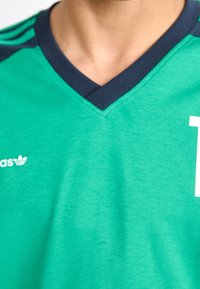 Lähikuva henkilöstä, joka pitää päällään vihreää urheiluopiskelijaa, jossa on tummansininen kaulus ja Adidas-logo vasemmalla puolella.