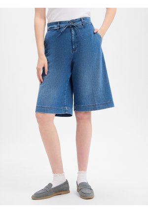 Personne portant un short en jean bleu de longueur moyenne avec une ceinture nouée, des chaussettes blanches et des chaussures grises sans lacets, debout avec une main dans la poche.