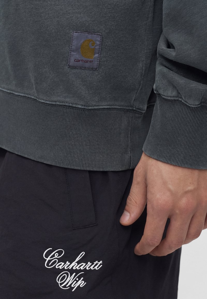 Gray Carhartt sweatshirt with logo patch above black pants featuring white "Carhartt Wip" embroidery, hand resting beside pants pocket.