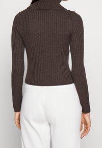 Brun ribbad tröja med hög krage, figurnära design, långa ärmar och texturerat mönster. Sömmen slutar vid midjan, i kombination med vita byxor.