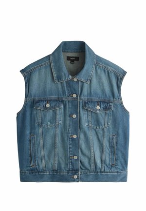 Junge Frau mit langem geflochtenem Haar trägt ärmellose Jeansweste, passenden Jeansrock, schwarze Keilsandalen und hält eine kleine dunkelbraune Handtasche.