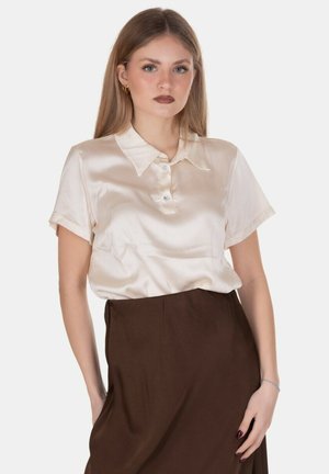 Giovane donna con lunghi capelli biondi che indossa una camicetta corta in raso color crema e una gonna marrone a vita alta, che posa su sfondo bianco.