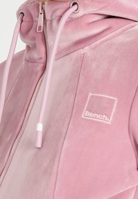 Mjuk rosa huvtröja med framdragkedja, dragsko och broderad Bench-logotyp på bröstet.